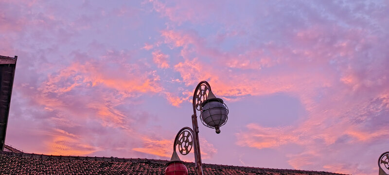 Evening sky with soft pink and purple clouds illuminated by the setting sun, seen over a historic building with an ornate street lamp - Powered by Adobe