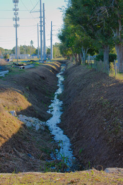 Rural Drainage Ditch With Water Channel, Power Lines, And Lined Trees Along Utility Corridor. 
A narrow earthen drainage ditch runs between grassy banks, bordered by a row of mature trees. Tall power 