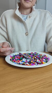 ​A woman sitting at a table uses a spoon to eat colorful plastic fuse beads from a white plate, symbolizing the ingestion of microplastics and the danger of endocrine disruptors