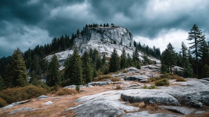 A dramatic mountain landscape featuring a large, light gray rock face rising dramatically under a stormy sky, with a dense forest of evergreens at its base.