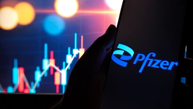 Hand holding smartphone displaying pfizer logo against backdrop of stock market graph