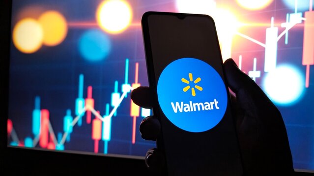 Hand holding smartphone displaying walmart logo against a background of stock market charts and graphs