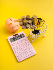 Creative financial success concept with piggy bank, coins, calculator showing profit, and golden trophy in cart, representing saving money, income growth, and sales rewards on yellow background.