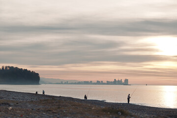 Beach at dusk with calm water and silhouettes of anglers along the shoreline, warm sky and distant skyline, tranquil mood and gentle waves reflecting sunset light over pebbled shore