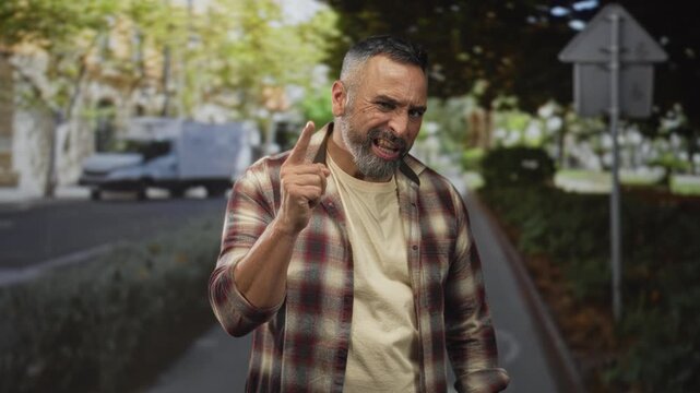 Man points finger to temple on street wearing plaid shirt and beard, grimacing with raised brow and clenched jaw; anger.