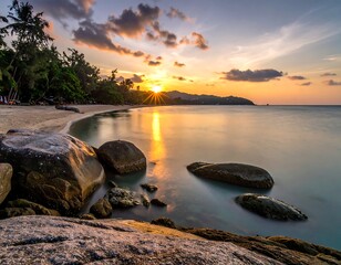 Beautiful sunset over a tropical beach. Golden light reflects on the calm water, with rocks in foreground and palm trees