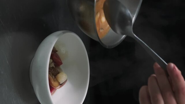 Vertical video. Cooking hot meal. Seafood soup. Chef pouring spoon steaming liquid in bowl with fresh tomatoes. Appetizing octopus clams culinary.