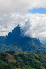 Obraz premium Dense white clouds drift across a dramatic, rugged mountain peak rising above lush green hills in the countryside between Vang Vieng and Phou Khoun, Laos. The scene features deep shadows, textured