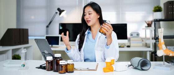 Professional asian female doctor in white gown sitting at desk with laptop, showing medicine bottle and explaining prescription during online consultation in hospital office