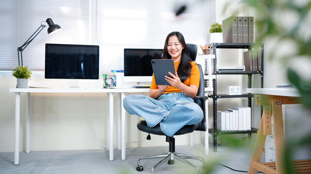 A young woman sitting comfortably cross-legged in a modern office chair, holding a tablet. Focus on flexible work and technology.