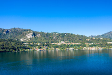 Fototapeta premium Panoramic view of hillside settlements and lush green forests reflecting on the calm blue waters of Lake Orta in northern Italy. The scene features rugged rocky slopes, scattered houses, and a vivid