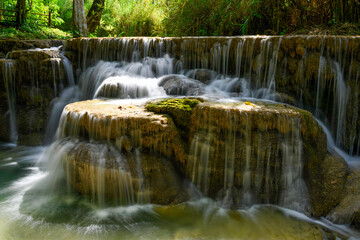 Soft sunlight highlights tiered limestone formations as water cascades gently over mossy rocks at Kuang Si Falls in northern Laos. The scene features lush green surroundings and tranquil flowing water © Florent