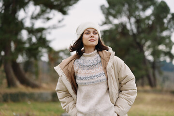 Woman in a beige coat and knit sweater stands outdoors in a park, wearing a knit beanie, conveying...
