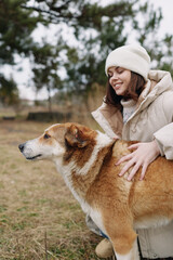 Woman in winter coat and knit hat smiles as she pets a brown and white dog outdoors in a park,...