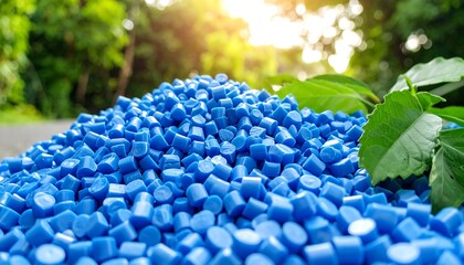 A close-up shot of a vibrant heap of blue plastic pellets. Green foliage and sunlight create a natural, textured backdrop
