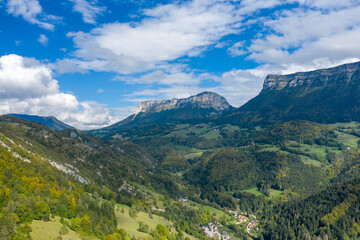 Fototapeta premium Wide aerial perspective of the Chartreuse Massif reveals dramatic limestone cliffs, rolling green valleys, and scattered forested slopes under a vibrant blue sky with scattered clouds. The scene