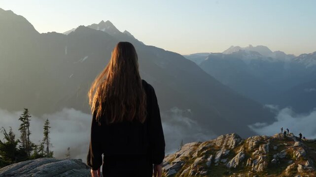 Camera follows young woman walking along a rocky slope. She stops, turns, and smiles toward the camera as clouds fill the valley behind her. Real-time sunrise golden hour footage.