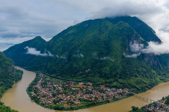 Aerial view of Nong Khiaw village along the muddy Nam Ou River, framed by steep green mountains and drifting low clouds in northern Laos. The scene features dense tropical forest, a prominent bridge