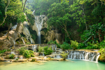 Multi-tiered cascades flow over pale limestone rocks surrounded by dense green foliage at Kuang Si Waterfalls in northern Laos. Sunlight filters through the tropical canopy, illuminating turquoise © Florent