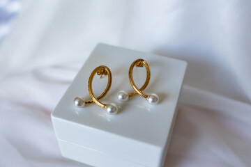 A pair of gold earrings with pearls on them are sitting on a white box. The pearls are small and the earrings are shaped like a fish. The earrings are the main focus of the image.