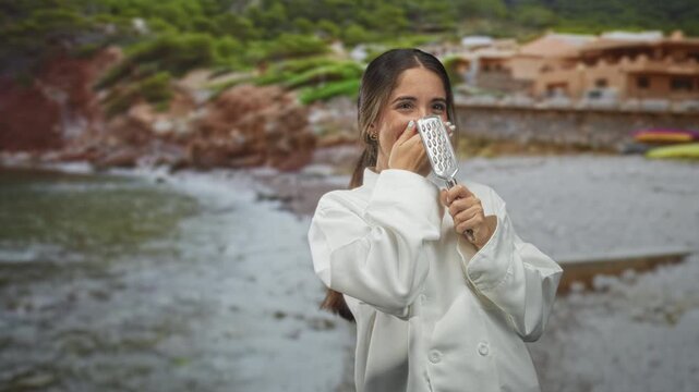 Woman chef in white jacket holds a grater, hand covers mouth laughing on a rocky beach by seaside houses; joy.