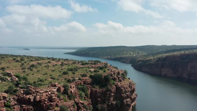 Expansive aerial showcases Gandikota Canyon with Pennar River reservoir curving through red cliffs, green plateaus, distant hills, and scattered vegetation under partly cloudy skies.