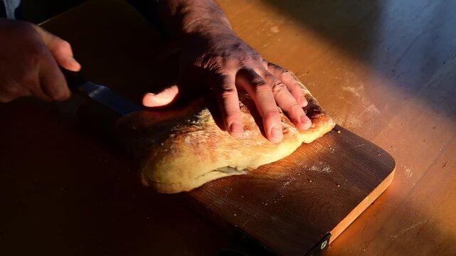 Close-up of male hand slicing fresh focaccia with knife to reveal airy interior, sunny day with warm golden light