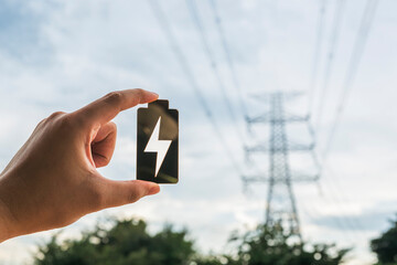 Hand holding battery icon with lightning symbol in front of power transmission tower and sky...