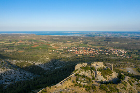 Ancient stone remains of Chateau de Opoul Perillos overlook a mosaic of farmland and a distant village, with the blue sea shimmering on the horizon under clear skies.