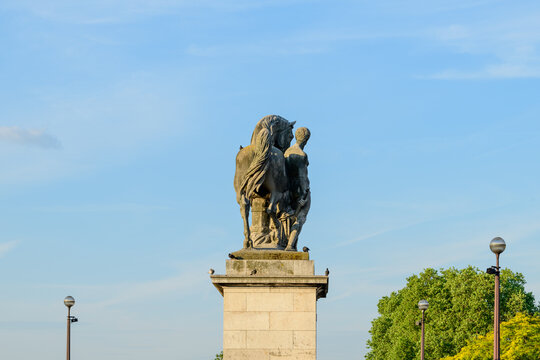 Backlit stone sculpture of a man and horse atop a pedestal on Pont de Iena in Paris, surrounded by blue sky and greenery in soft evening light.