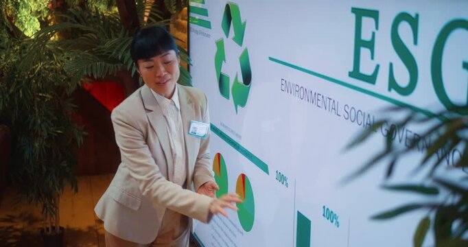 Japanese businesswoman explains ESG data and sustainability strategy, gesturing toward charts and recycling symbol on large digital display during corporate presentation in modern eco office.