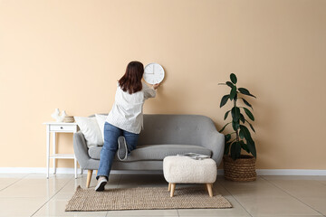 Young woman hanging clock on beige wall in living room, back view