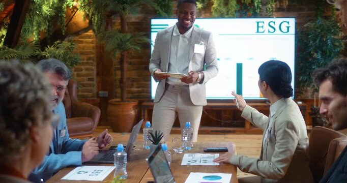 Business team laughs openly during ESG meeting as Black businessman leads presentation, creating positive and relaxed atmosphere while discussing sustainability strategy in modern corporate office.