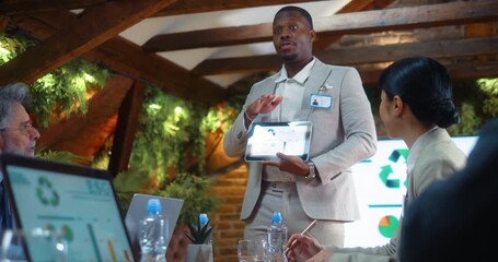 Black businessman presents ESG data on tablet to corporate team, explaining charts and analytics while leading discussion during business meeting in modern eco office environment. - Powered by Adobe