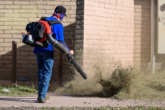 Worker using a backpack leaf blower to clear weeds along a brick wall, representing grounds maintenance, landscaping services, and outdoor cleaning operations