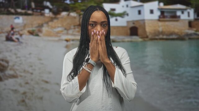 Black woman with long braids covers her mouth with hands, wearing white blouse and bracelets, standing by seaside building while smiling; surprise wonder.