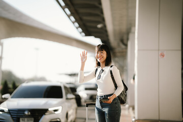 Happy young Asian woman traveler waving her hand while holding a smartphone and suitcase at an airport curbside, waiting for pickup or transportation, representing modern travel lifestyle.