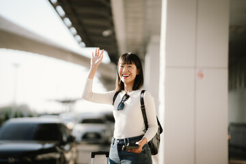 Happy young Asian woman traveler waving her hand while holding a smartphone and suitcase at an airport curbside, waiting for pickup or transportation, representing modern travel lifestyle.