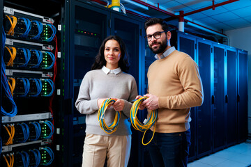 Caucasian young adult woman and Caucasian young adult man standing in server room holding network cables, both looking at camera, surrounded by data center equipment and server racks