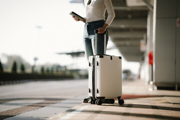 Young Asian woman traveler standing at an airport curbside with a rolling suitcase and smartphone, waiting for pickup or transportation, representing modern travel and urban mobility. © Witoon