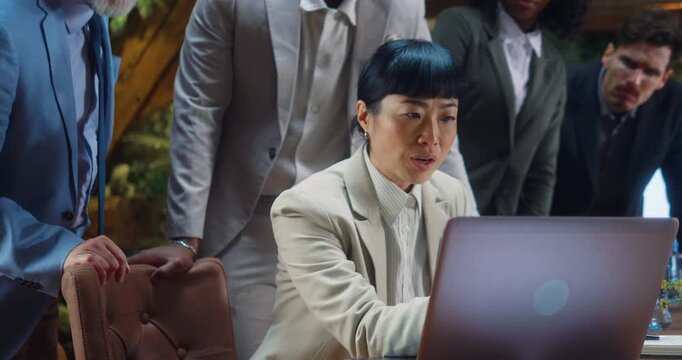 Japanese businesswoman reads good news from laptop as team reacts happily, celebrating success, cooperation and shared victory during multicultural business meeting in modern office.