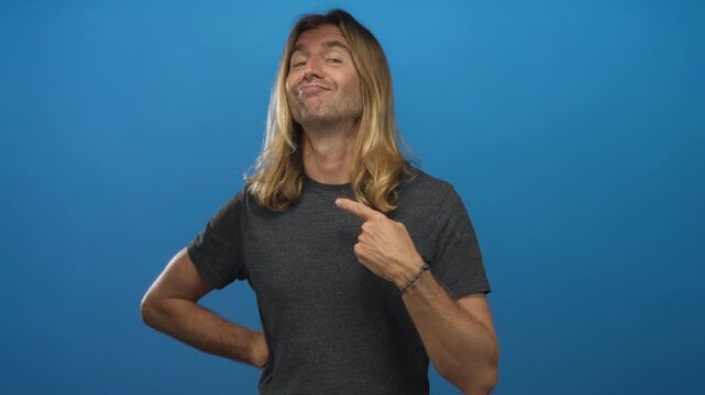 Blond man with long hair wearing grey t shirt pointing finger to chest with one hand on hip in studio with blue backdrop; confidence.