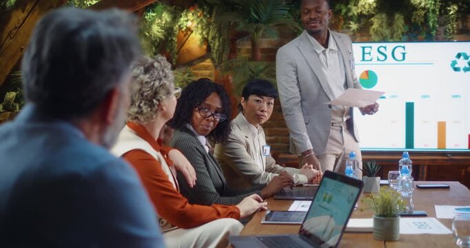 Business meeting with active discussion as senior colleague raises hand to ask question while Black businessman leads presentation in modern eco conference room.