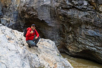 Happy asian male hiker stand on a light colored rock beside a gently flowing stream, holding a sturdy hiking stick inside the national park.