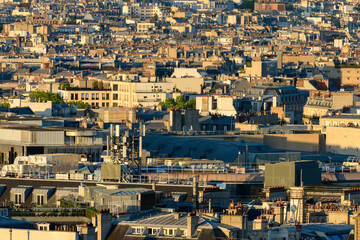 Fototapeta premium Dense layers of Paris rooftops with varied architectural styles and rooftop structures, illuminated by warm evening light. The urban landscape features a rhythmic pattern of chimneys, antennas, and