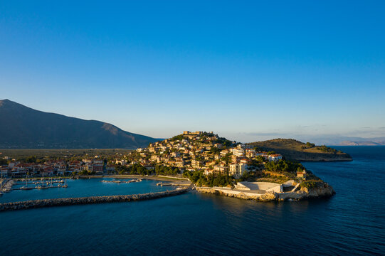 Picturesque coastal town of Paralio Astros with clustered houses on a hilltop, a small marina, and deep blue sea under clear evening light. The tranquil scene features warm sunlight, calm water, and a