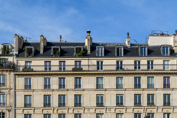 Obraz premium Elegant Haussmannian apartment facade on Ile Saint-Louis in Paris, featuring cream stone walls, wrought iron balconies, and a row of dormer windows under a blue sky.