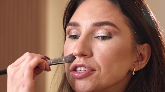 Professional makeup artist applying blush, lipstick and fixing spray to smiling woman in modern beauty salon. Close up shots of eyeshadow palette, brush on eyelids, warm golden colors and wooden inter