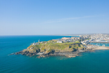 Wide-angle drone photo captures Pointe du Roc's grassy headland and dramatic rocky shoreline meeting clear blue sea, with the town and marina in the distance under bright sunlight. The landscape is