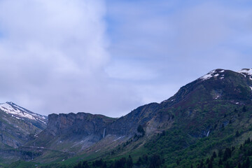 Fototapeta premium Multiple waterfalls stream down steep rocky cliffs and green slopes, with patches of snow and a wide expanse of moody sky above in the Roselend area of the French Alps.